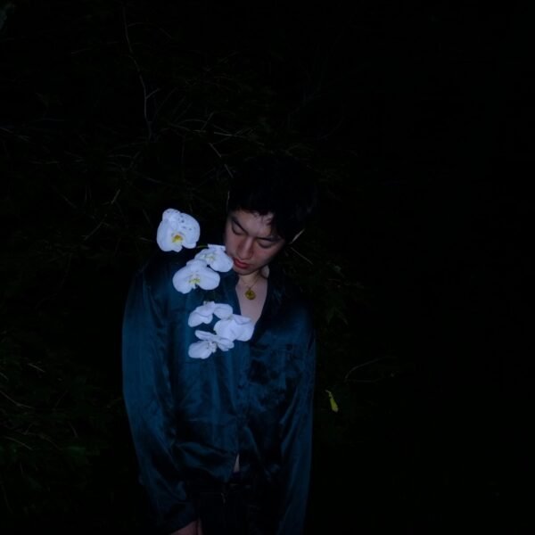A man stands in darkness, adorned with delicate white flowers on his shirt, creating a moody and stylish portrait.
